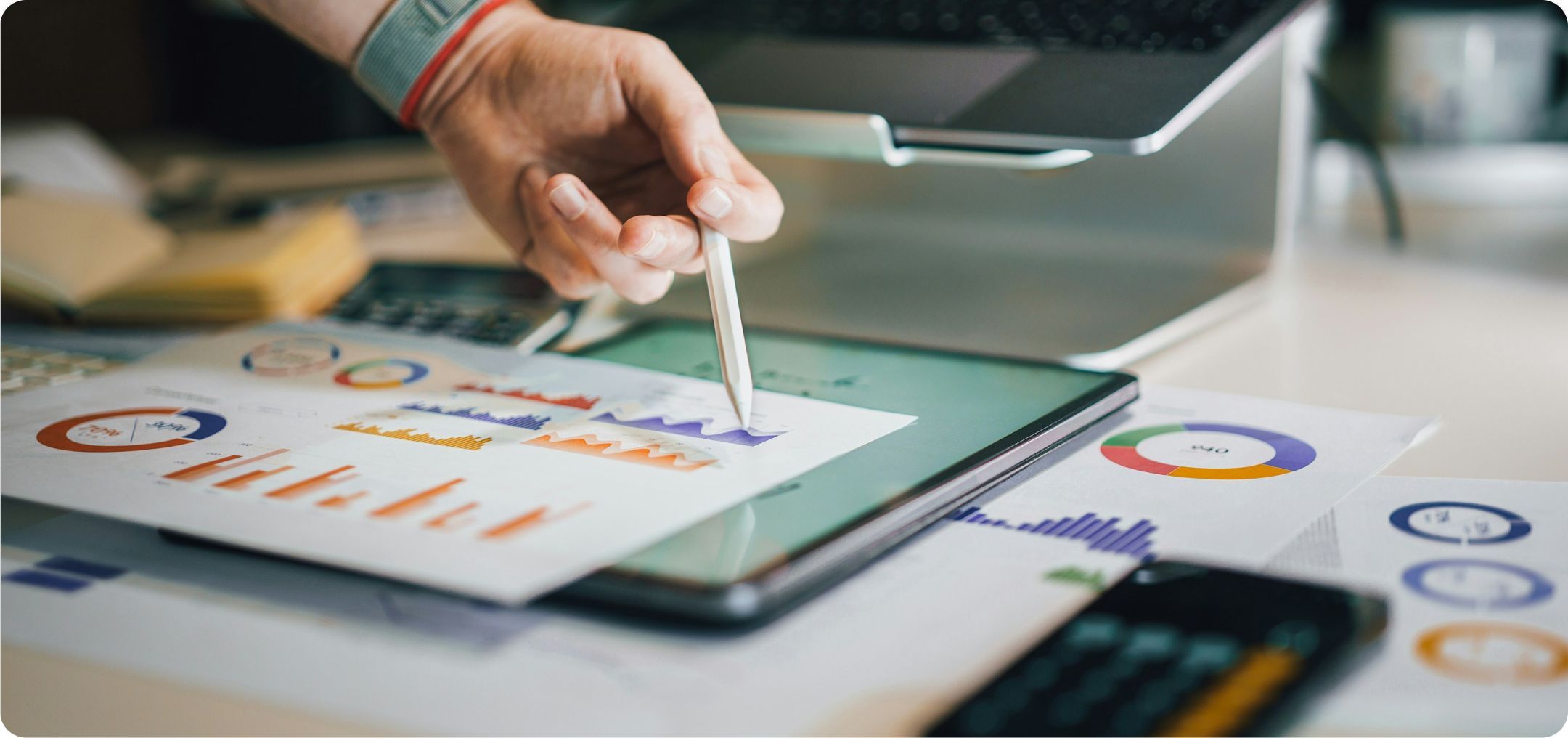 A hand pointing to charts on a page that is on a desk full of charts and graphs. Representing data analytics techniques. 